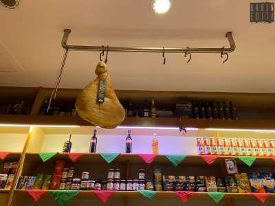 Sullo sfondo si stagliano mensole di vari generi alimentari, mentre da una trave sul soffitto pende una coscia di prosciutto crudo